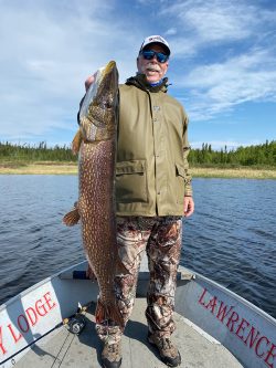 A man standing on a boat holds up a large fish he caught, smiling at the camera on a clear day.