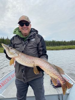 Man in outdoor clothing holding a large fish on a boat with water and trees in the background.