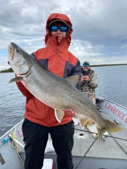 Person in a red jacket holding a large fish on a boat, with another person taking a photo in the background.