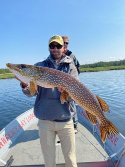 Person holding a large northern pike fish on a boat, with another person standing behind, on a lake under a clear sky.