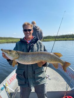 A person standing on a boat holds a large fish, with another person fishing in the background on a sunny day.