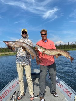 Two men standing on a boat each holding a large fish, with water and trees visible in the background.
