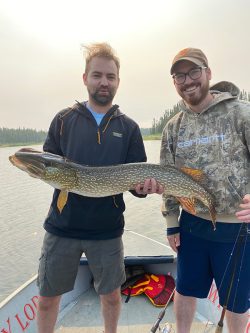 Two men on a boat hold a large fish together, with water and trees visible in the background.