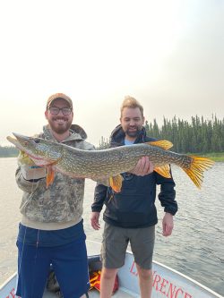 Two men on a boat, one holding a large fish, with trees and water in the background.