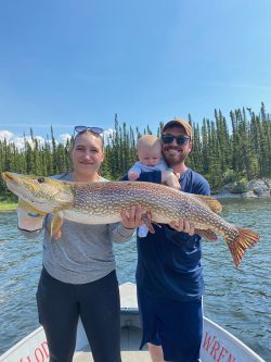A woman and man holding a large fish with a baby on a boat, trees and water in the background.