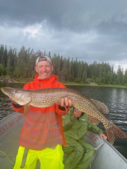 Person standing on a boat holding a large fish with a lake and forest in the background under a cloudy sky.
