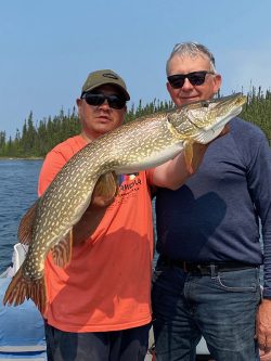 Two men standing on a boat hold a large fish with a lake and pine trees visible in the background.