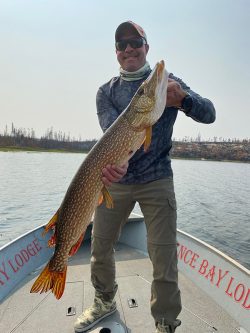Person standing on a boat holding a large fish with two hands, water and trees visible in the background.
