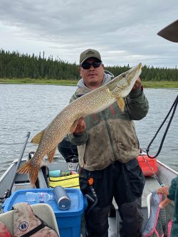 A man in a boat holds a large northern pike fish with a lake and forest in the background.