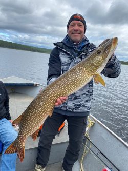 Man standing in a boat holds a large northern pike fish, with a lake and trees visible in the background.