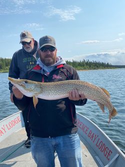 Two men on a boat, one holding a large fish, with a forest and lake in the background.
