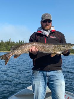 Man standing on a boat holding a large fish over the water, wearing sunglasses, a hat, and a black jacket.