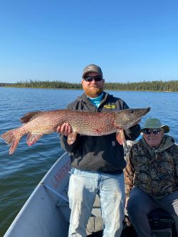 A man standing on a boat holds a large fish, while another man sits beside him; lake and trees in the background.