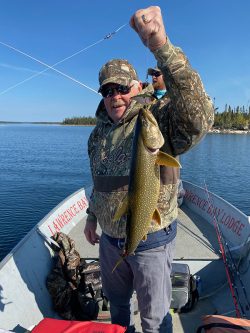 Person in camouflage clothing holding a large fish on a boat labeled "Lawrence Bay Lodge" on a lake.