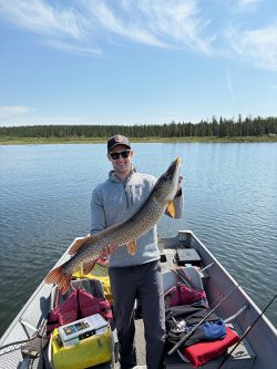 Person standing on a boat holds a large fish with both hands, surrounded by fishing gear on a calm lake.