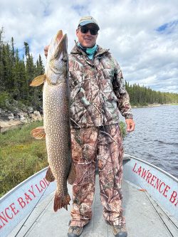 Person in camouflage clothing standing on a boat holds a large fish next to a lake with trees in the background.