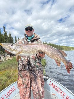 Person in camouflage gear holding a large fish on a boat, with trees and a lake in the background under a cloudy sky.