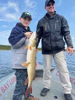 A boy holding a large fish stands next to an adult man on a boat on a lake, both smiling at the camera.