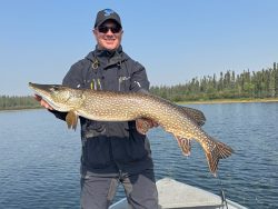 Man standing on a boat holds a large northern pike fish with a lake and trees in the background.