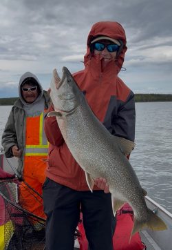 Person in a red jacket holds a large fish on a boat; another person in an orange vest stands behind them.