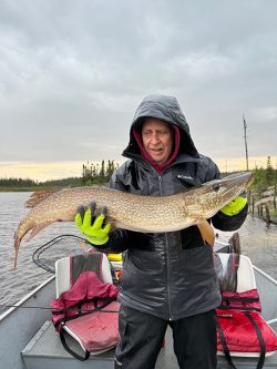 Person standing on a boat holding a large fish with both hands, wearing a raincoat and neon gloves.