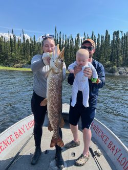 Two adults on a boat hold a large fish and a baby, with water and a forested shore in the background.