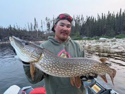 Person holding a large northern pike fish on a boat, with a lake and forested shoreline in the background.