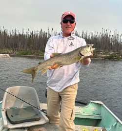 Man standing in a small boat holds a large fish with both hands; fishing rod rests on the boat seat nearby.