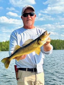 Man wearing sunglasses holds a large fish while standing on a boat in a lake, with trees and sky in the background.
