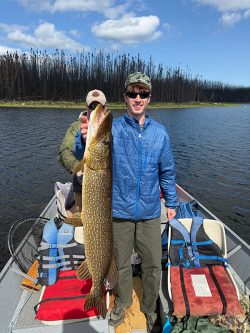 Man standing on a boat holds up a large fish; another person sits behind him. Lake and burned trees in the background.