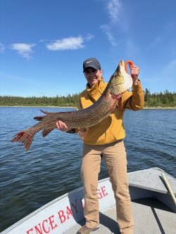 Person standing on a boat holding a large fish with water and trees visible in the background.