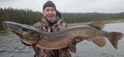 Man in camo jacket and black beanie holding a large northern pike fish by the water with trees in the background.
