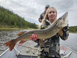 Woman in camouflage jacket holding a large fish on a boat, with another person and trees in the background.