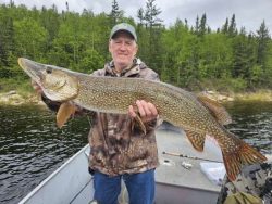 Man standing on a boat holding a large northern pike with a forested shoreline in the background.