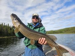 Person in outdoor gear holding a large fish on a boat, with a lake and trees in the background.