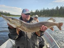 Man holding a large northern pike fish on a boat, with another person and trees in the background.