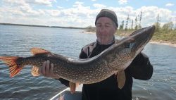Person holding a large northern pike fish on a boat with a lake and trees in the background.