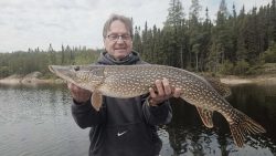 A man stands by a lake holding a large northern pike fish with both hands, trees visible in the background.