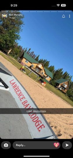 A sandy beach with a sign reading "LAWRENCE BAY LODGE," log cabins, and trees under a clear blue sky.