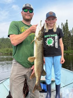 An adult and a child on a boat hold up a large fish with trees and water in the background.