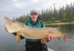 Older man in a green jacket holds up a large fish on a boat with trees and water in the background.