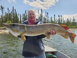 Person holding a large northern pike fish on a boat in a lake, with trees and a partly cloudy sky in the background.