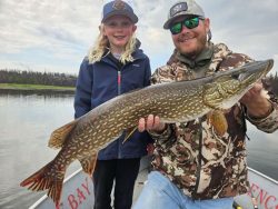 An adult and child on a boat display a large fish they caught, with calm water and trees in the background.