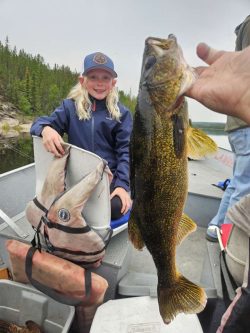 A person holds up a large fish on a boat while a smiling child sits nearby with a life jacket.
