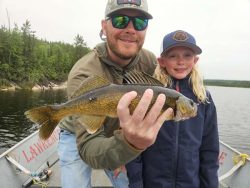 An adult and child on a boat hold up a freshly caught fish with a lake and forest in the background.
