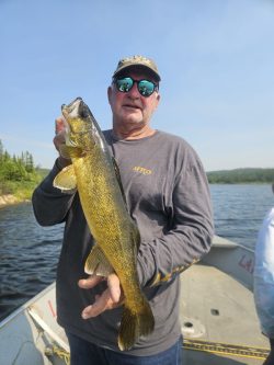 Man standing on a boat holding a large fish with one hand, with a lake and trees in the background.