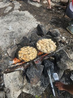 Two pans of battered fish frying over an open campfire on a grill, with rocks and people visible nearby.