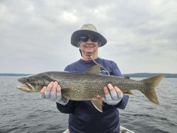 Person wearing hat and sunglasses holds a large fish on a boat with a lake and cloudy sky in the background.
