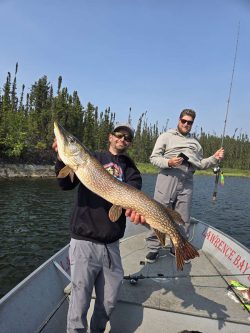 Two people stand in a boat on a lake; one holds a large fish, and the other holds a fishing rod.