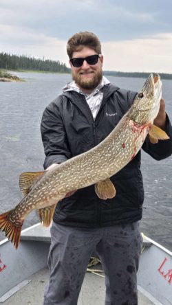 A man wearing sunglasses holds a large fish on a boat with water and trees in the background.